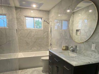 Remodeled bathroom with marble-look porcelain tile, a tiled tub-shower and a round mirror over a dark vanity