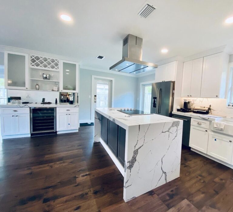 Remodeled kitchen with white shaker cabinets, a dark gray island wrapped in a marble-look quartz waterfall top, an induction cooktop under a stainless island hood, a farmhouse sink and a bar wall with a wine fridge
