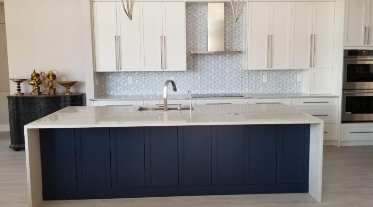 Kitchen with a navy shaker island under a white quartz waterfall top, white shaker wall cabinets, a geometric tile backsplash, a stainless chimney hood and double wall ovens