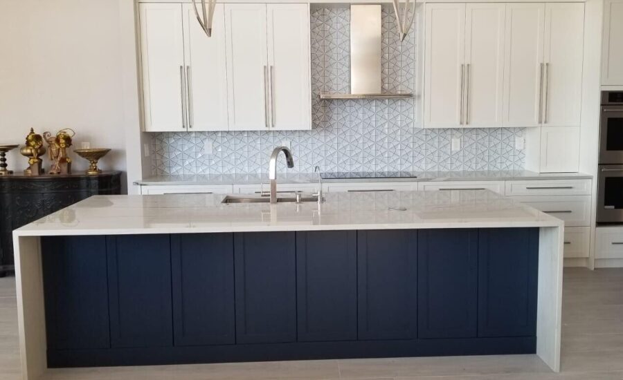 Kitchen with a navy shaker island under a white quartz waterfall top, white shaker wall cabinets, a geometric tile backsplash, a stainless chimney hood and double wall ovens