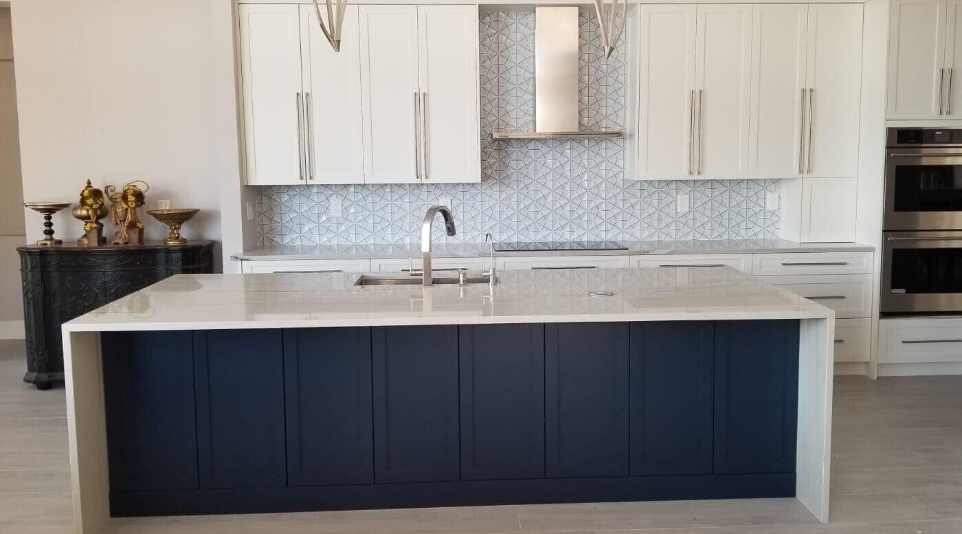 Kitchen with a navy shaker island under a white quartz waterfall top, white shaker wall cabinets, a geometric tile backsplash, a stainless chimney hood and double wall ovens